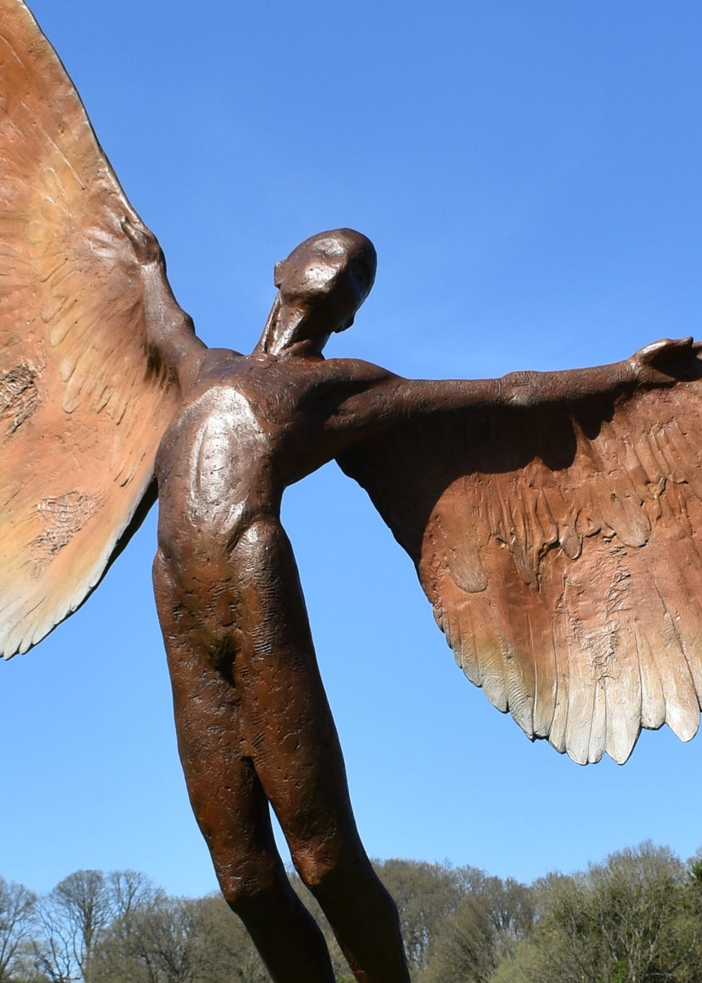 Icarus I — monumental bronze sculpture by Nicola Godden, depicting a stylised male figure with immense outstretched wings, leaning forward as if preparing for flight. Rich bronze patina with warm coppery browns fading into lighter golden tips across the feathered wings. Originally featured at the London 2012 Olympic Village and sold at Sotheby’s. Set outdoors against a clear blue sky and classical architectural backdrop.