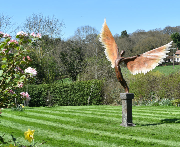 Icarus I — monumental bronze sculpture by Nicola Godden, depicting a stylised male figure with immense outstretched wings, leaning forward as if preparing for flight. Rich bronze patina with warm coppery browns fading into lighter golden tips across the feathered wings. Originally featured at the London 2012 Olympic Village and sold at Sotheby’s. Set outdoors against a clear blue sky and classical architectural backdrop.