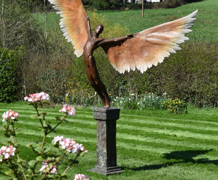 Icarus I — monumental bronze sculpture by Nicola Godden, depicting a stylised male figure with immense outstretched wings, leaning forward as if preparing for flight. Rich bronze patina with warm coppery browns fading into lighter golden tips across the feathered wings. Originally featured at the London 2012 Olympic Village and sold at Sotheby’s. Set outdoors against a clear blue sky and classical architectural backdrop.