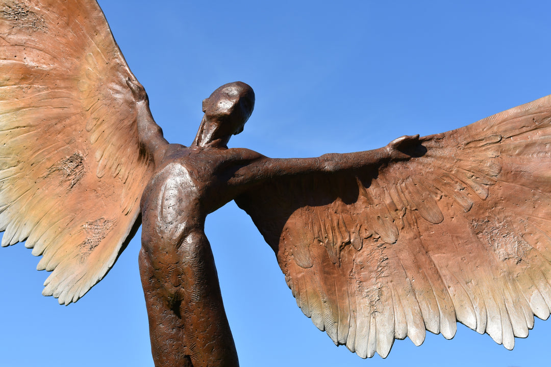 Icarus I — monumental bronze sculpture by Nicola Godden, depicting a stylised male figure with immense outstretched wings, leaning forward as if preparing for flight. Rich bronze patina with warm coppery browns fading into lighter golden tips across the feathered wings. Originally featured at the London 2012 Olympic Village and sold at Sotheby’s. Set outdoors against a clear blue sky and classical architectural backdrop.