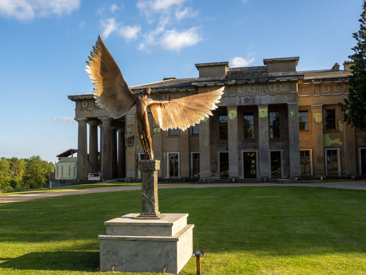Icarus I — monumental bronze sculpture by Nicola Godden, depicting a stylised male figure with immense outstretched wings, leaning forward as if preparing for flight. Rich bronze patina with warm coppery browns fading into lighter golden tips across the feathered wings. Originally featured at the London 2012 Olympic Village and sold at Sotheby’s. Set outdoors against a clear blue sky and classical architectural backdrop.