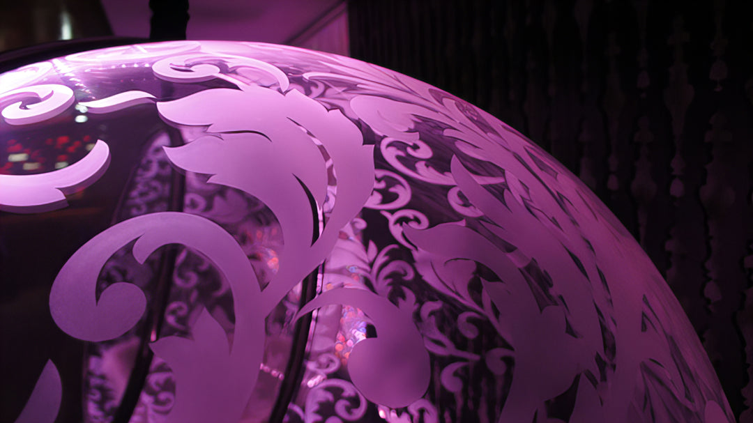 “Illuminated Bubble Chair – Flock Pattern” by Ben Rousseau – a glowing hanging chair crafted from stainless steel, engraved acrylic, and RGB LED light. The spherical design emits soft pink and violet tones, lined with a Mongolian sheepskin cushion.

