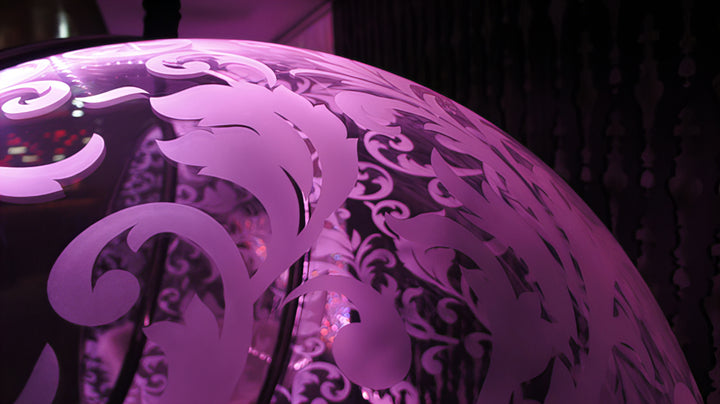 “Illuminated Bubble Chair – Flock Pattern” by Ben Rousseau – a glowing hanging chair crafted from stainless steel, engraved acrylic, and RGB LED light. The spherical design emits soft pink and violet tones, lined with a Mongolian sheepskin cushion.

