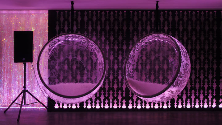 “Illuminated Bubble Chair – Flock Pattern” by Ben Rousseau – a glowing hanging chair crafted from stainless steel, engraved acrylic, and RGB LED light. The spherical design emits soft pink and violet tones, lined with a Mongolian sheepskin cushion.

