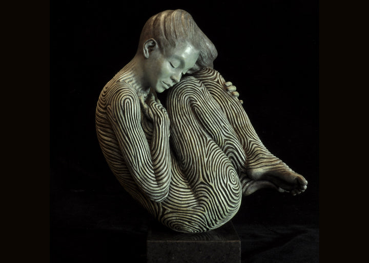 Bronze sculpture of a female figure curled into a foetal position, her body entirely covered in flowing fingerprint-like whorls, loops and arches. The surface is painted in tones of grey, charcoal and soft white, creating a tactile pattern that highlights the contours of the form. The figure’s expression is peaceful and introspective, with hair sweeping gently to one side. Set outdoors against soft greenery, the sculpture conveys calm, protection and individuality.