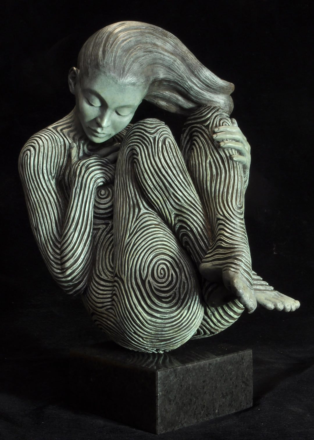 Bronze sculpture of a female figure curled into a foetal position, her body entirely covered in flowing fingerprint-like whorls, loops and arches. The surface is painted in tones of grey, charcoal and soft white, creating a tactile pattern that highlights the contours of the form. The figure’s expression is peaceful and introspective, with hair sweeping gently to one side. Set outdoors against soft greenery, the sculpture conveys calm, protection and individuality.