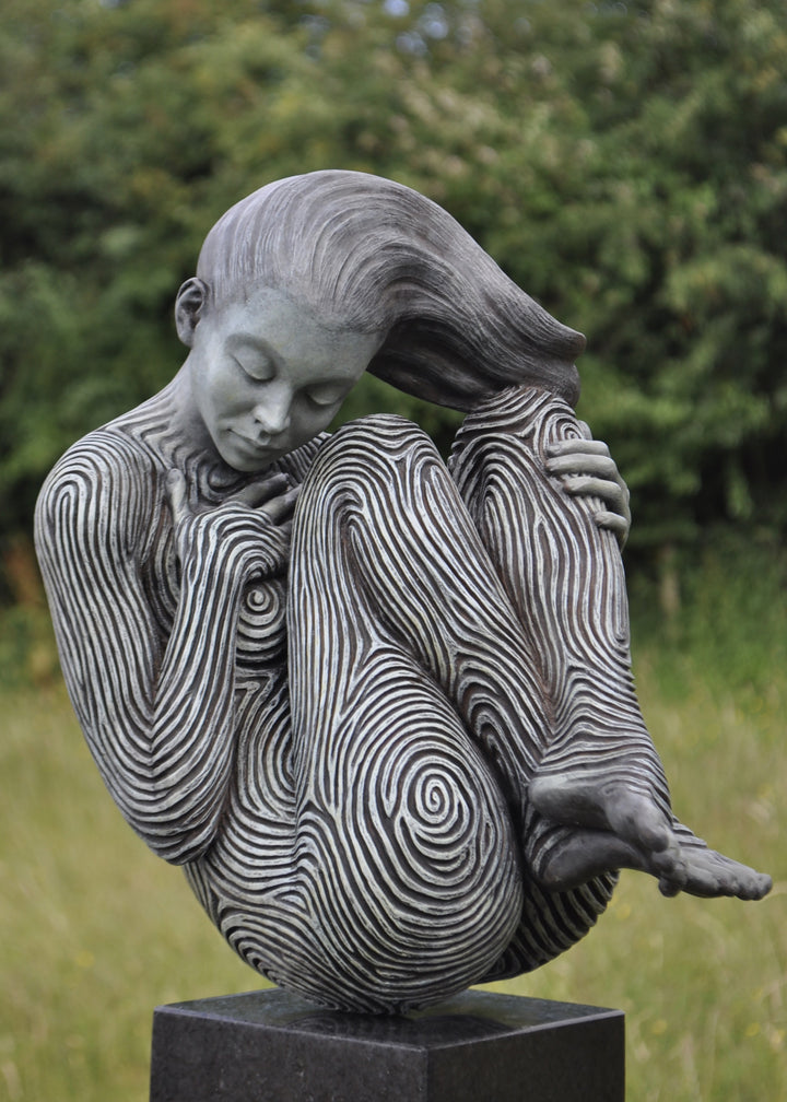 Bronze sculpture of a female figure curled into a foetal position, her body entirely covered in flowing fingerprint-like whorls, loops and arches. The surface is painted in tones of grey, charcoal and soft white, creating a tactile pattern that highlights the contours of the form. The figure’s expression is peaceful and introspective, with hair sweeping gently to one side. Set outdoors against soft greenery, the sculpture conveys calm, protection and individuality.