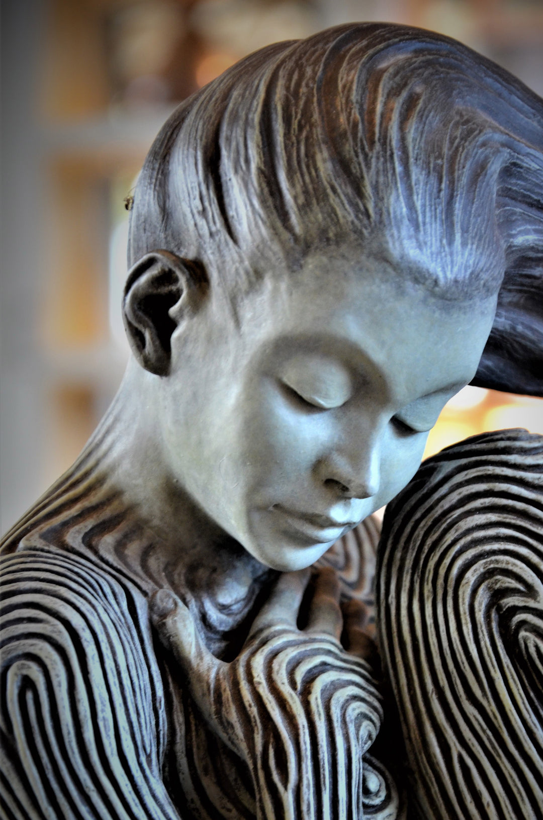 Bronze sculpture of a female figure curled into a foetal position, her body entirely covered in flowing fingerprint-like whorls, loops and arches. The surface is painted in tones of grey, charcoal and soft white, creating a tactile pattern that highlights the contours of the form. The figure’s expression is peaceful and introspective, with hair sweeping gently to one side. Set outdoors against soft greenery, the sculpture conveys calm, protection and individuality.