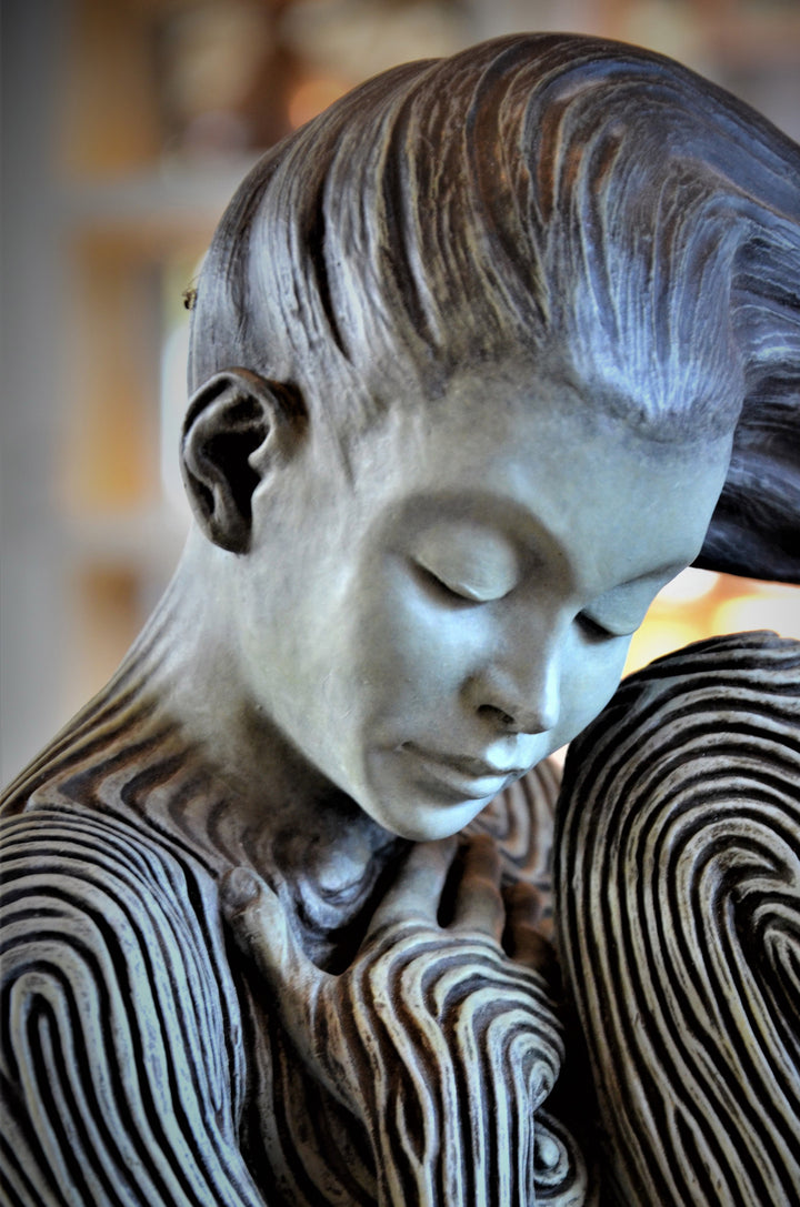Bronze sculpture of a female figure curled into a foetal position, her body entirely covered in flowing fingerprint-like whorls, loops and arches. The surface is painted in tones of grey, charcoal and soft white, creating a tactile pattern that highlights the contours of the form. The figure’s expression is peaceful and introspective, with hair sweeping gently to one side. Set outdoors against soft greenery, the sculpture conveys calm, protection and individuality.