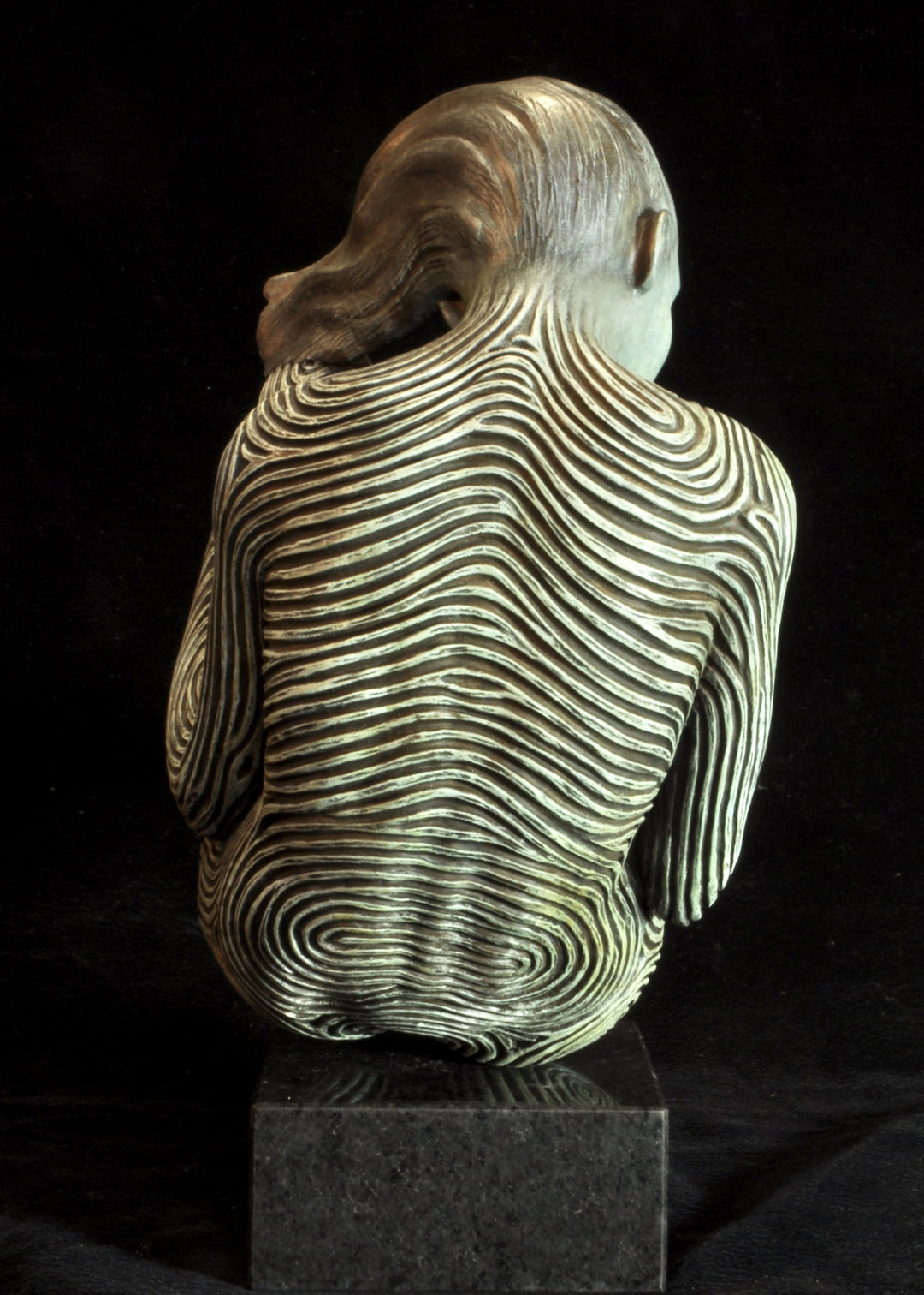 Bronze sculpture of a female figure curled into a foetal position, her body entirely covered in flowing fingerprint-like whorls, loops and arches. The surface is painted in tones of grey, charcoal and soft white, creating a tactile pattern that highlights the contours of the form. The figure’s expression is peaceful and introspective, with hair sweeping gently to one side. Set outdoors against soft greenery, the sculpture conveys calm, protection and individuality.