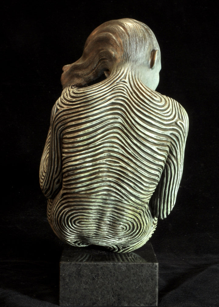 Bronze sculpture of a female figure curled into a foetal position, her body entirely covered in flowing fingerprint-like whorls, loops and arches. The surface is painted in tones of grey, charcoal and soft white, creating a tactile pattern that highlights the contours of the form. The figure’s expression is peaceful and introspective, with hair sweeping gently to one side. Set outdoors against soft greenery, the sculpture conveys calm, protection and individuality.