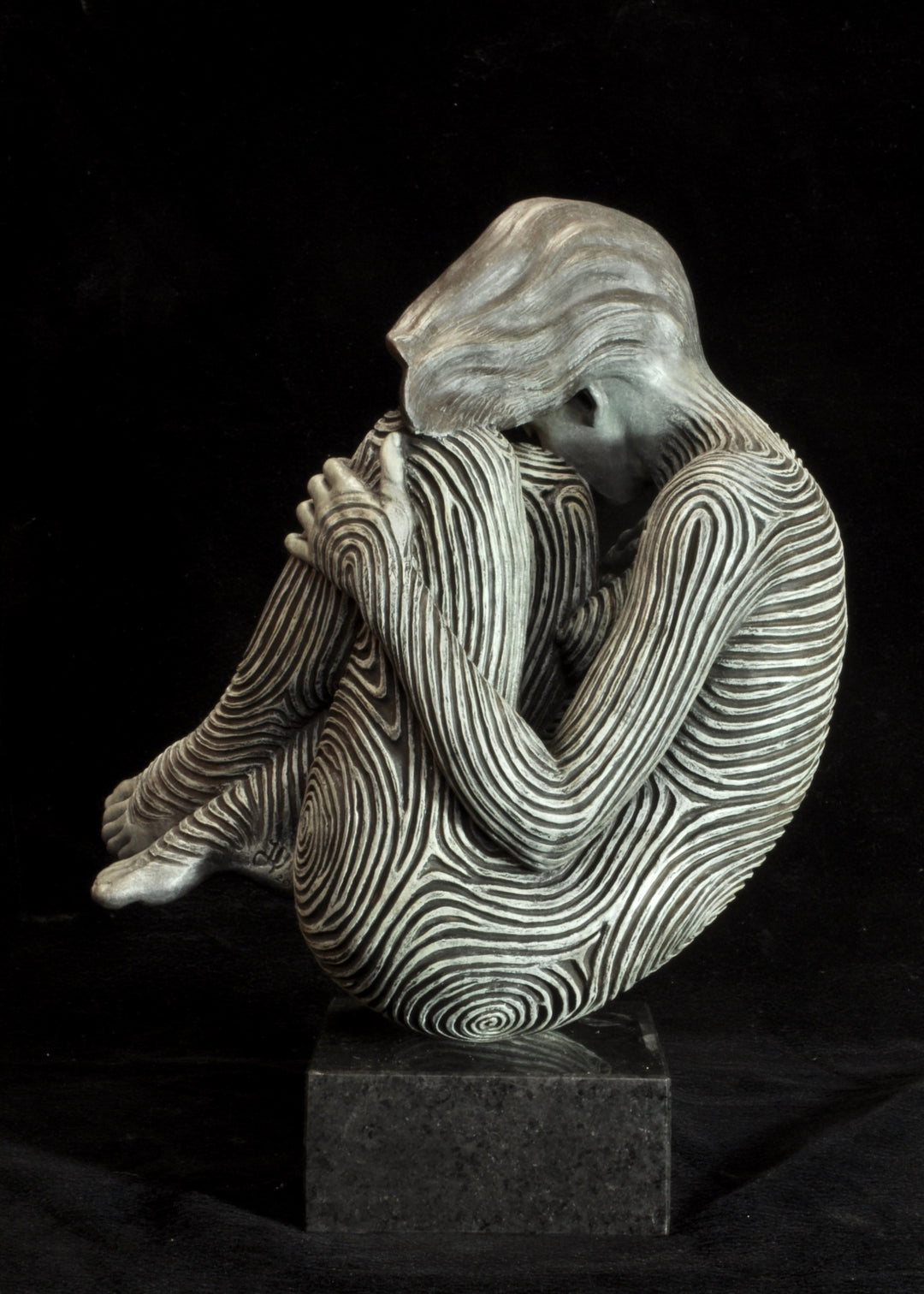 Bronze sculpture of a female figure curled into a foetal position, her body entirely covered in flowing fingerprint-like whorls, loops and arches. The surface is painted in tones of grey, charcoal and soft white, creating a tactile pattern that highlights the contours of the form. The figure’s expression is peaceful and introspective, with hair sweeping gently to one side. Set outdoors against soft greenery, the sculpture conveys calm, protection and individuality.