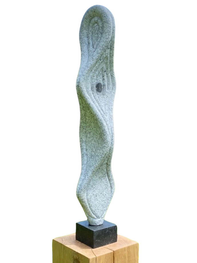 Abstract sculpture In the Mind’s Eye – 2024 by Hamish Holman, carved from Cornish granite into a tall, twisting vertical form with flowing contours and textured surface, mounted on a black stone base and oak plinth, displayed outdoors in a meadow setting.