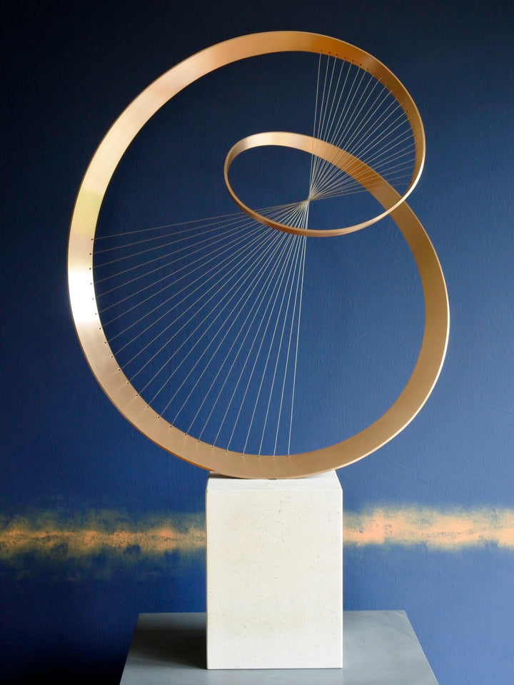 Fabricated bronze sculpture titled Infinity by Thomas Joynes, mounted on a Portland stone plinth. The work features two intersecting bronze curves connected by fine stainless steel micro wires, forming a minimal yet elegant spatial tension. The clean, modernist structure appears weightless and musical, its elliptical ribbons casting delicate linear shadows, evoking motion and balance. Photographed against a deep blue backdrop.