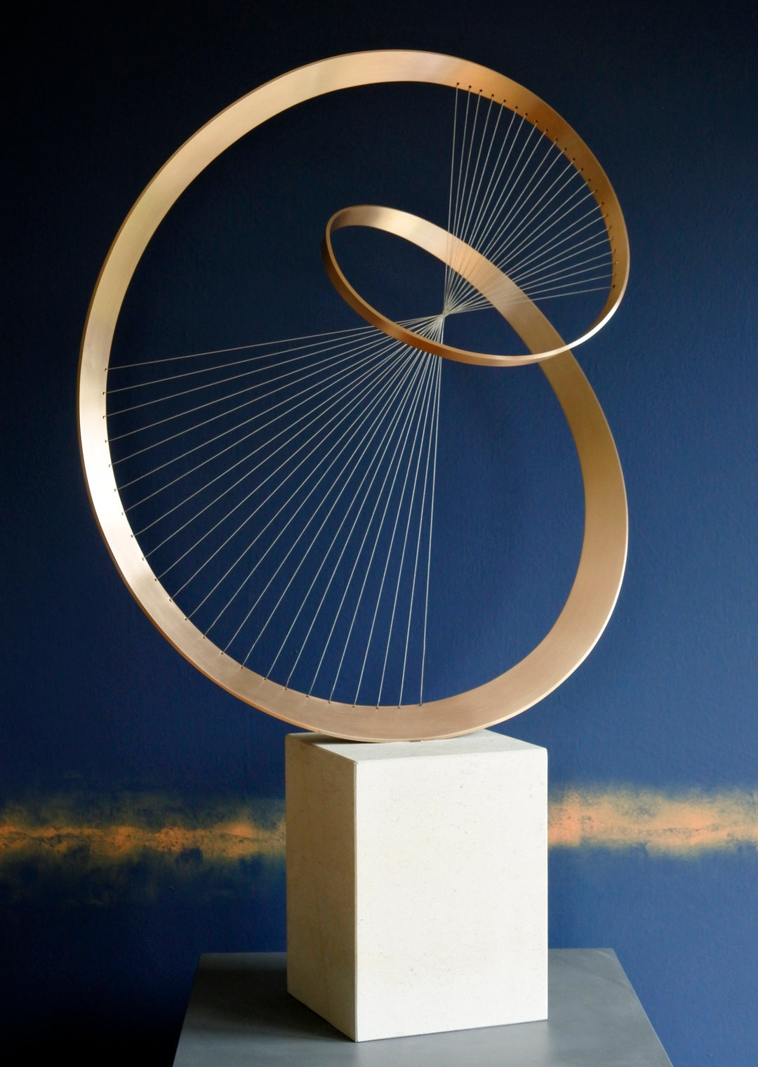 Fabricated bronze sculpture titled Infinity by Thomas Joynes, mounted on a Portland stone plinth. The work features two intersecting bronze curves connected by fine stainless steel micro wires, forming a minimal yet elegant spatial tension. The clean, modernist structure appears weightless and musical, its elliptical ribbons casting delicate linear shadows, evoking motion and balance. Photographed against a deep blue backdrop.