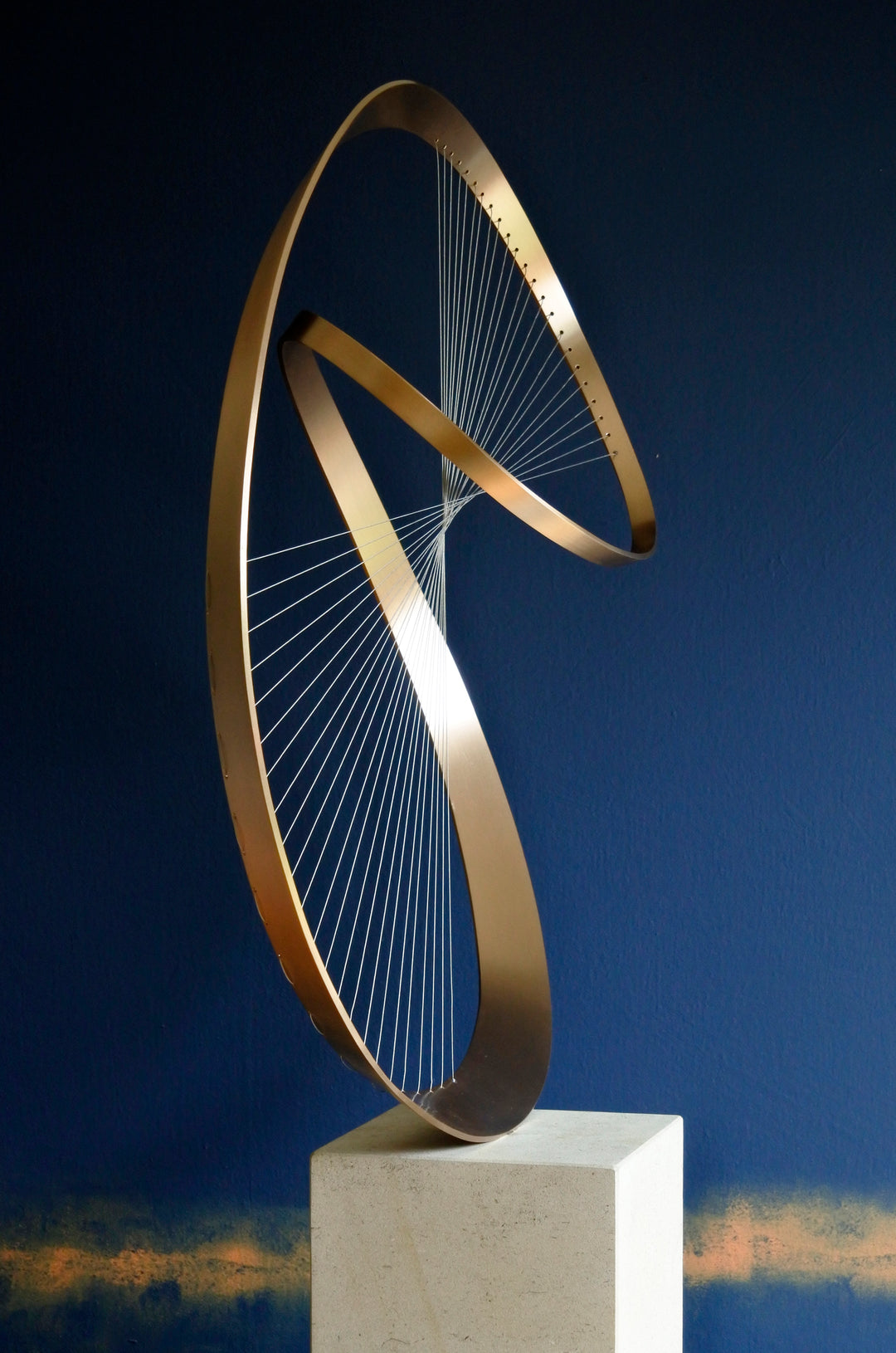 Fabricated bronze sculpture titled Infinity by Thomas Joynes, mounted on a Portland stone plinth. The work features two intersecting bronze curves connected by fine stainless steel micro wires, forming a minimal yet elegant spatial tension. The clean, modernist structure appears weightless and musical, its elliptical ribbons casting delicate linear shadows, evoking motion and balance. Photographed against a deep blue backdrop.