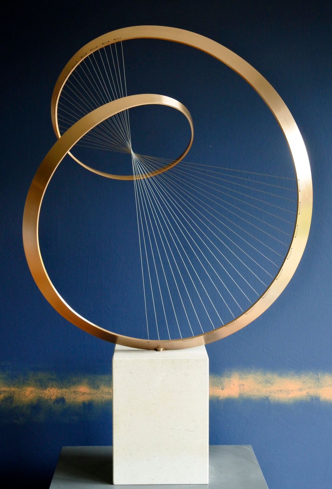 Fabricated bronze sculpture titled Infinity by Thomas Joynes, mounted on a Portland stone plinth. The work features two intersecting bronze curves connected by fine stainless steel micro wires, forming a minimal yet elegant spatial tension. The clean, modernist structure appears weightless and musical, its elliptical ribbons casting delicate linear shadows, evoking motion and balance. Photographed against a deep blue backdrop.