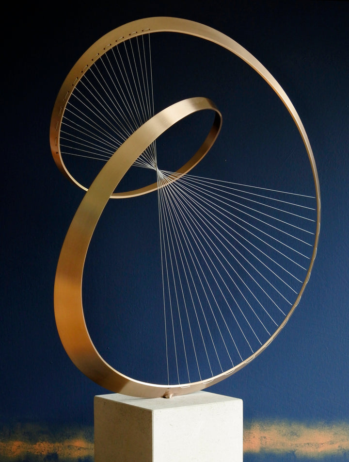 Fabricated bronze sculpture titled Infinity by Thomas Joynes, mounted on a Portland stone plinth. The work features two intersecting bronze curves connected by fine stainless steel micro wires, forming a minimal yet elegant spatial tension. The clean, modernist structure appears weightless and musical, its elliptical ribbons casting delicate linear shadows, evoking motion and balance. Photographed against a deep blue backdrop.