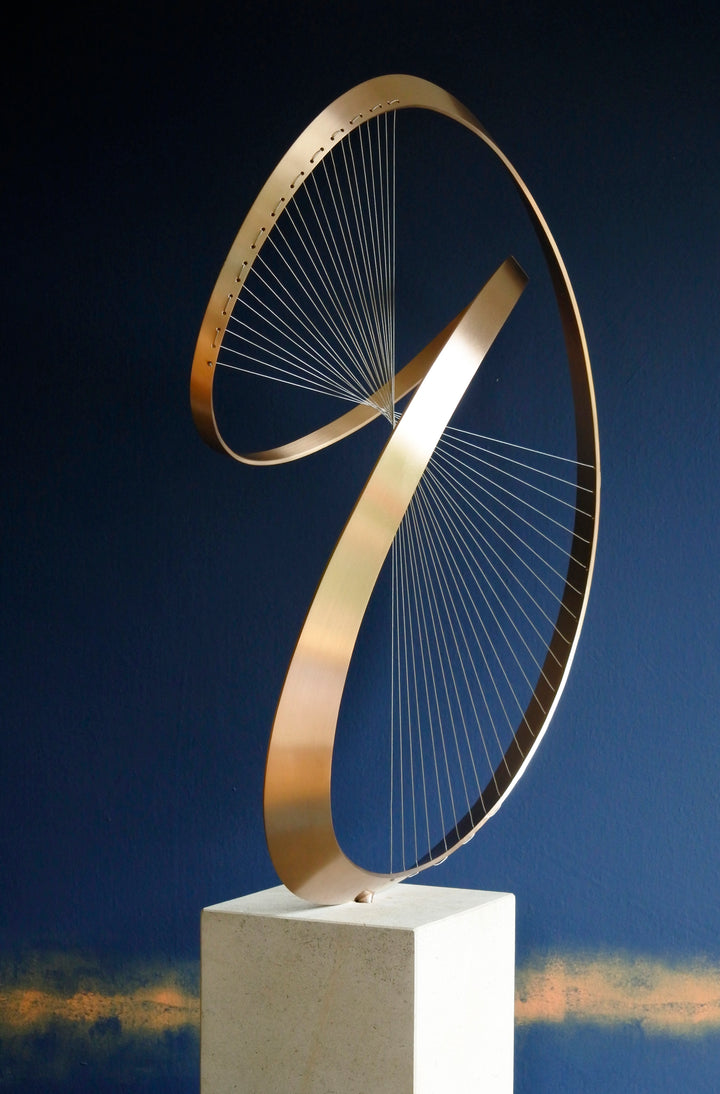 Fabricated bronze sculpture titled Infinity by Thomas Joynes, mounted on a Portland stone plinth. The work features two intersecting bronze curves connected by fine stainless steel micro wires, forming a minimal yet elegant spatial tension. The clean, modernist structure appears weightless and musical, its elliptical ribbons casting delicate linear shadows, evoking motion and balance. Photographed against a deep blue backdrop.
