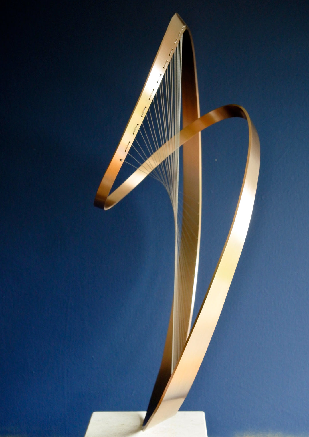 Fabricated bronze sculpture titled Infinity by Thomas Joynes, mounted on a Portland stone plinth. The work features two intersecting bronze curves connected by fine stainless steel micro wires, forming a minimal yet elegant spatial tension. The clean, modernist structure appears weightless and musical, its elliptical ribbons casting delicate linear shadows, evoking motion and balance. Photographed against a deep blue backdrop.