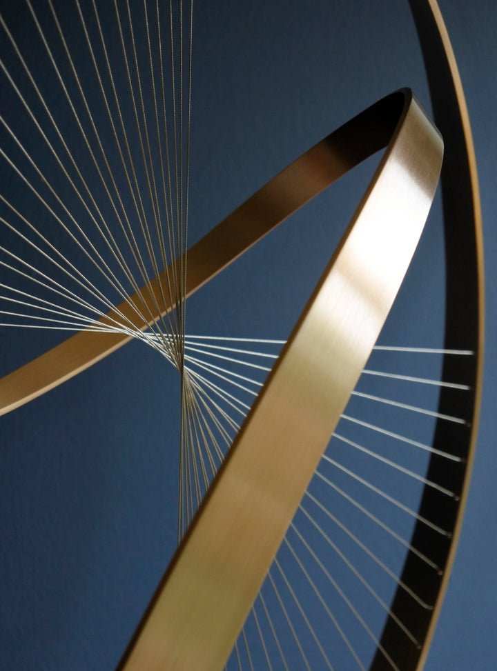 Fabricated bronze sculpture titled Infinity by Thomas Joynes, mounted on a Portland stone plinth. The work features two intersecting bronze curves connected by fine stainless steel micro wires, forming a minimal yet elegant spatial tension. The clean, modernist structure appears weightless and musical, its elliptical ribbons casting delicate linear shadows, evoking motion and balance. Photographed against a deep blue backdrop.