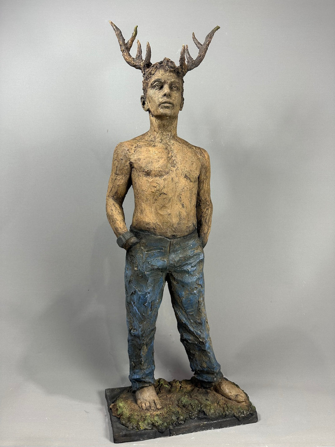Contemporary ceramic full-figure sculpture by Eve Shepherd depicting a standing male figure with antlers, merging human and animal forms, featuring raw hand-built clay textures, bare torso, distressed blue trousers and an earth-toned base, exploring instinct, identity and the tension between civilisation and primal nature