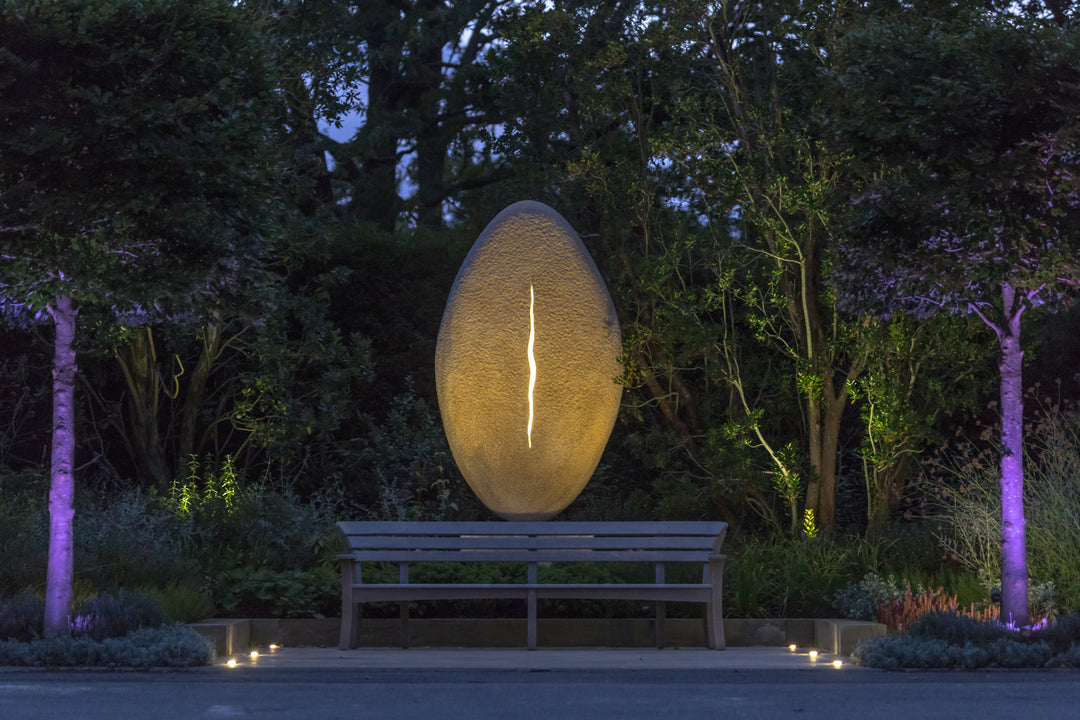 ‘New Beginnings’ by Nicolas Moreton MRSS, a unique commissioned contemporary abstract stone sculpture carved from Ancaster Weatherbed Limestone, featuring a tall, upright oval form with a textured surface and a narrow, irregular vertical opening illuminated by warm white LED light. The sculpture stands on a Portland limestone base in a landscaped garden at dusk, with its illuminated centre contrasting against the dark surrounding trees, purple garden lighting and evening sky.