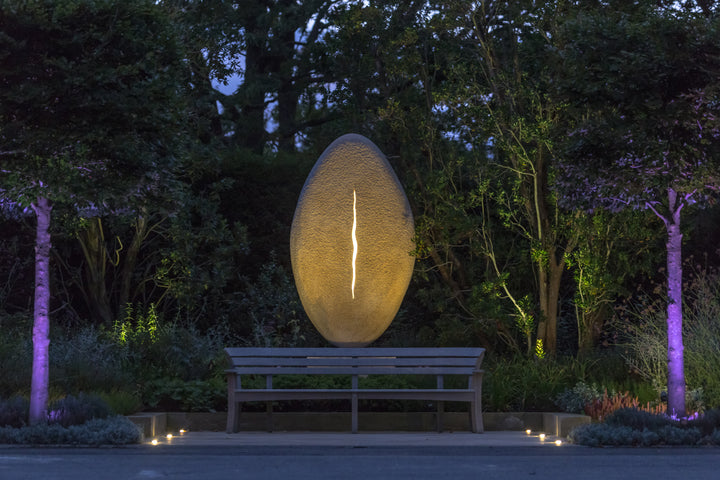 ‘New Beginnings’ by Nicolas Moreton MRSS, a unique commissioned contemporary abstract stone sculpture carved from Ancaster Weatherbed Limestone, featuring a tall, upright oval form with a textured surface and a narrow, irregular vertical opening illuminated by warm white LED light. The sculpture stands on a Portland limestone base in a landscaped garden at dusk, with its illuminated centre contrasting against the dark surrounding trees, purple garden lighting and evening sky.