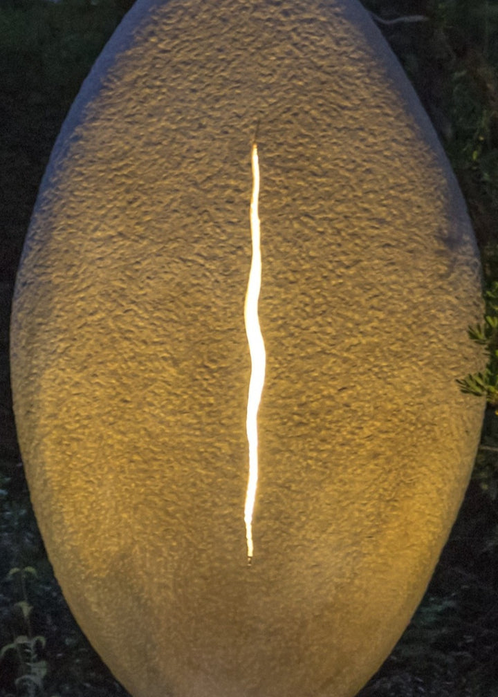 ‘New Beginnings’ by Nicolas Moreton MRSS, a unique commissioned contemporary abstract stone sculpture carved from Ancaster Weatherbed Limestone, featuring a tall, upright oval form with a textured surface and a narrow, irregular vertical opening illuminated by warm white LED light. The sculpture stands on a Portland limestone base in a landscaped garden at dusk, with its illuminated centre contrasting against the dark surrounding trees, purple garden lighting and evening sky.