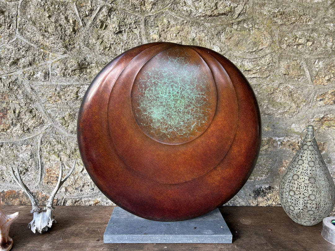 Circular bronze sculpture by Dominic Welch, Ionian Moon, with layered concentric curves and a patinated turquoise-green centre. The warm bronze surface contrasts with the cool central core, evoking lunar presence and timeless balance.