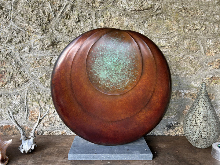 Circular bronze sculpture by Dominic Welch, Ionian Moon, with layered concentric curves and a patinated turquoise-green centre. The warm bronze surface contrasts with the cool central core, evoking lunar presence and timeless balance.
