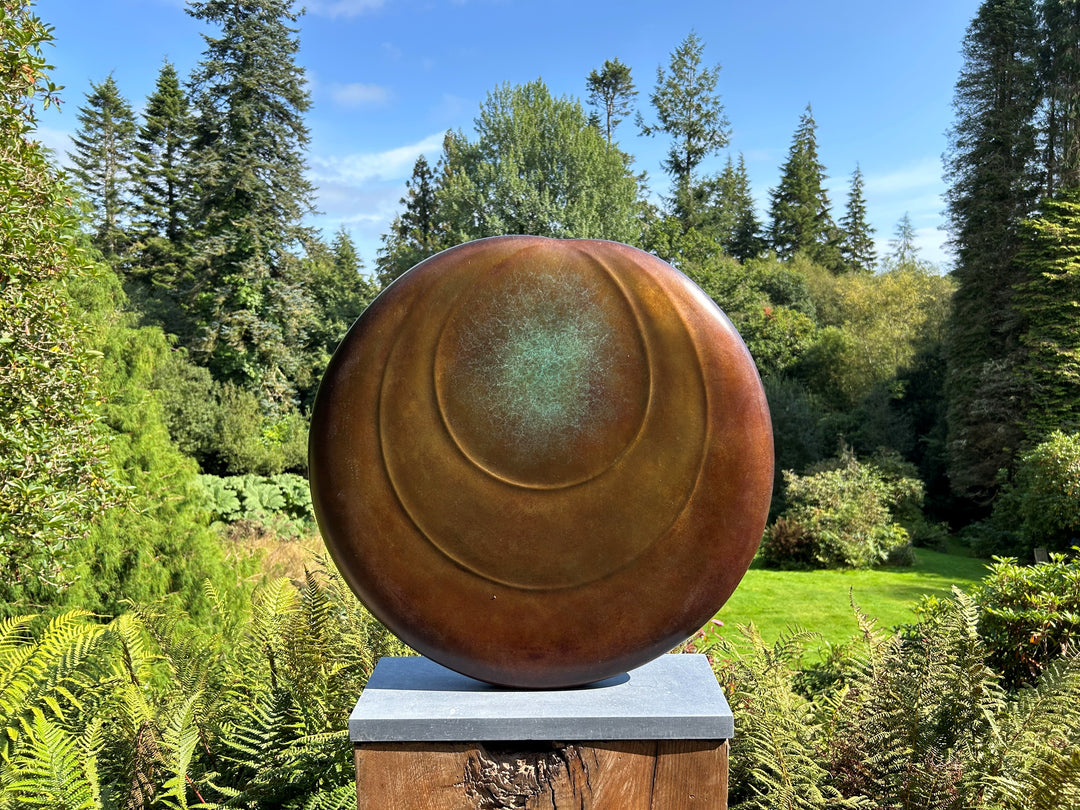 Circular bronze sculpture by Dominic Welch, Ionian Moon, with layered concentric curves and a patinated turquoise-green centre. The warm bronze surface contrasts with the cool central core, evoking lunar presence and timeless balance.