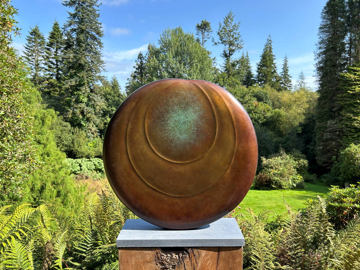 Circular bronze sculpture by Dominic Welch, Ionian Moon, with layered concentric curves and a patinated turquoise-green centre. The warm bronze surface contrasts with the cool central core, evoking lunar presence and timeless balance.
