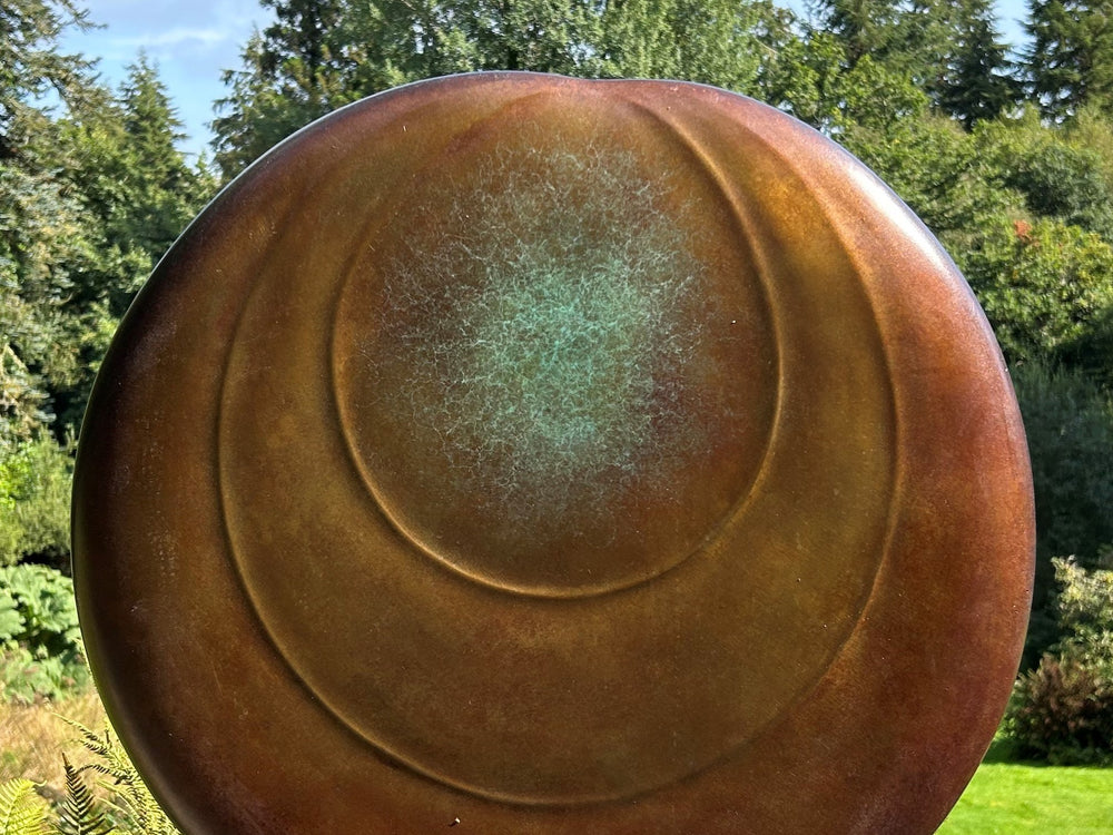 Circular bronze sculpture by Dominic Welch, Ionian Moon, with layered concentric curves and a patinated turquoise-green centre. The warm bronze surface contrasts with the cool central core, evoking lunar presence and timeless balance.