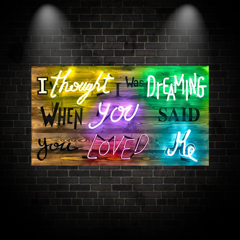 A multicoloured neon artwork by Matthew Bracey spelling out “I thought I was dreaming when you said you loved me” in a mix of script and block fonts. Each neon word glows in a different hue—yellow, green, white, purple, pink, and cyan—mounted against rustic, stained wood planks that transition in gradient colour tones. The vibrant piece blends romance, nostalgia, and urban edge with a playful pop-art twist.