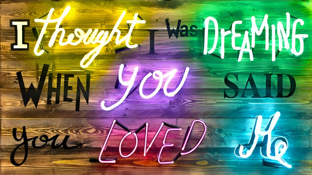 A multicoloured neon artwork by Matthew Bracey spelling out “I thought I was dreaming when you said you loved me” in a mix of script and block fonts. Each neon word glows in a different hue—yellow, green, white, purple, pink, and cyan—mounted against rustic, stained wood planks that transition in gradient colour tones. The vibrant piece blends romance, nostalgia, and urban edge with a playful pop-art twist.