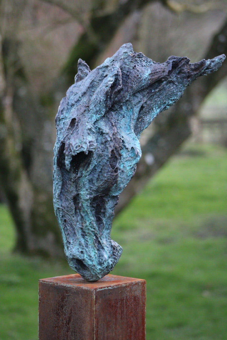 Abstract bronze sculpture of a horse’s head, mounted on a tall corten steel plinth, displayed in various garden and outdoor settings. The textured verdigris patina highlights the expressive contours and emotional form of the equine subject.