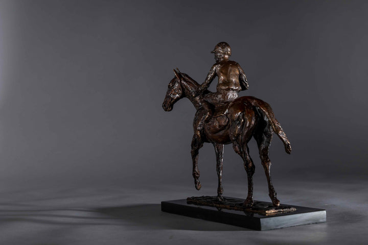 Bronze sculpture of a racehorse and jockey, titled Little White Lie, featuring a classic stance with refined detailing that captures both the poise of the rider and the strength of the thoroughbred. Mounted on a dark slate base, presented against a grey studio background.