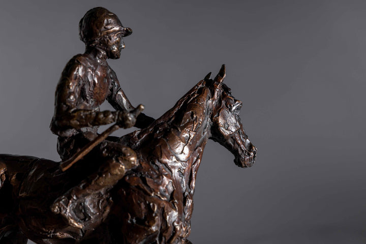 Bronze sculpture of a racehorse and jockey, titled Little White Lie, featuring a classic stance with refined detailing that captures both the poise of the rider and the strength of the thoroughbred. Mounted on a dark slate base, presented against a grey studio background.