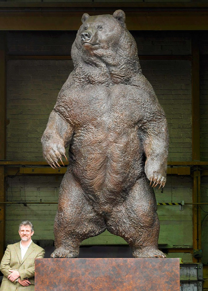 Monumental bronze sculpture of a life-size Kodiak brown bear standing upright on a large plinth inside an industrial foundry setting. The bear’s powerful muscular form, dense fur texture, and extended claws are rendered with extreme realism, conveying scale, strength, and dominance, while a human figure at the base provides dramatic contrast to emphasise the sculpture’s immense height and physical presence.