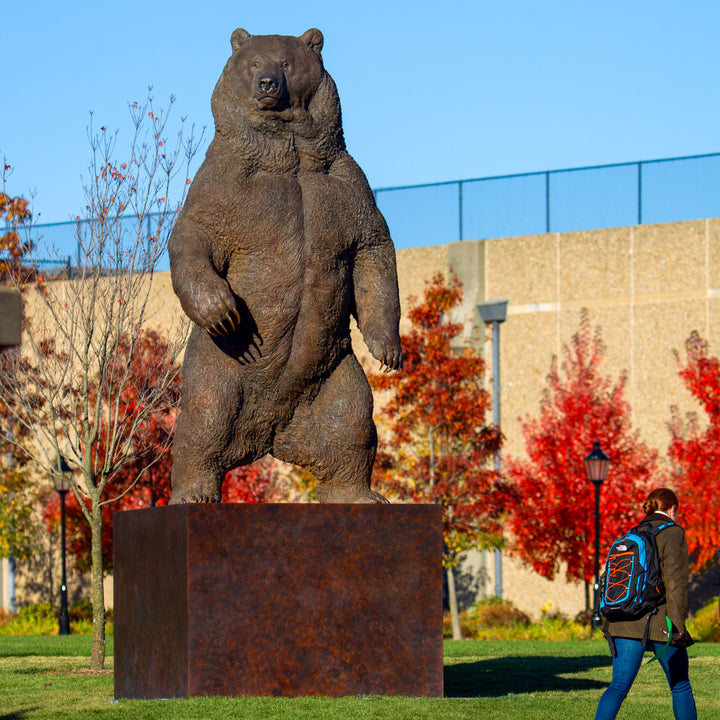 Monumental bronze sculpture of a life-size Kodiak brown bear standing upright on a large plinth inside an industrial foundry setting. The bear’s powerful muscular form, dense fur texture, and extended claws are rendered with extreme realism, conveying scale, strength, and dominance, while a human figure at the base provides dramatic contrast to emphasise the sculpture’s immense height and physical presence.