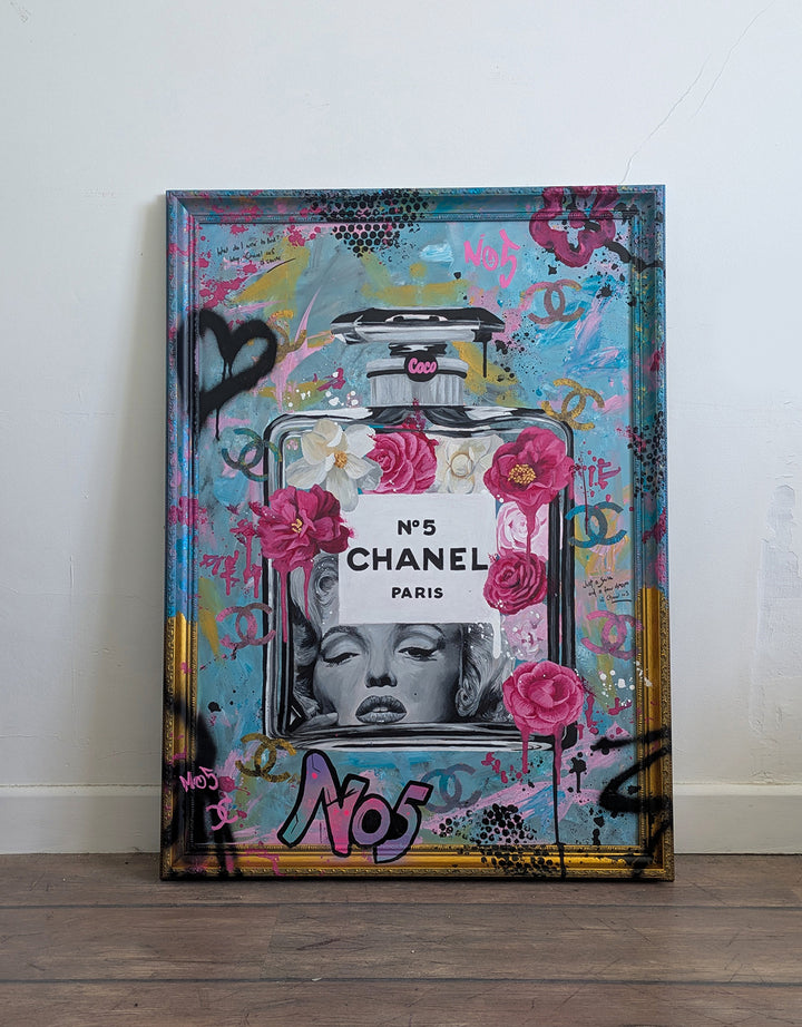 "Marilyn Monroe X Chanel No.5" – a bold embellished art print by Ian Salmon, featuring a black-and-white portrait of Marilyn Monroe embedded in a painted Chanel No.5 bottle, surrounded by vivid pink and red Camellia flowers, graffiti-style branding, and set against a turquoise, pastel blue, and gold accented background with a hand-painted custom frame.