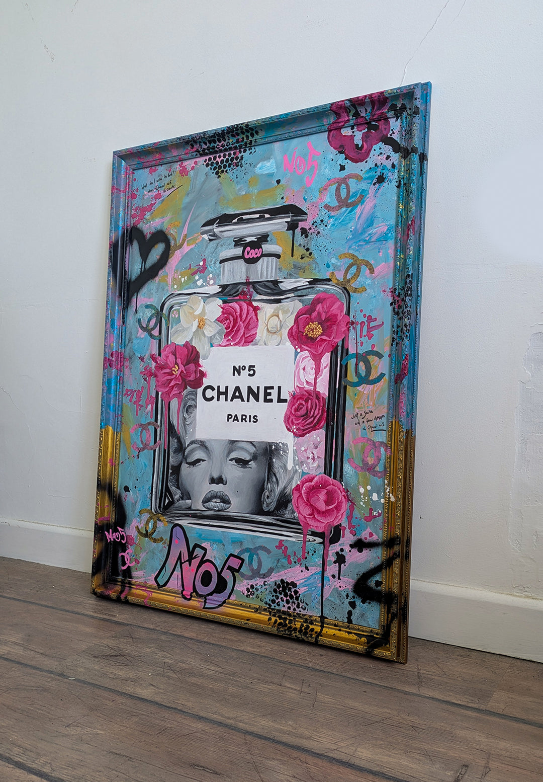 "Marilyn Monroe X Chanel No.5" – a bold embellished art print by Ian Salmon, featuring a black-and-white portrait of Marilyn Monroe embedded in a painted Chanel No.5 bottle, surrounded by vivid pink and red Camellia flowers, graffiti-style branding, and set against a turquoise, pastel blue, and gold accented background with a hand-painted custom frame.