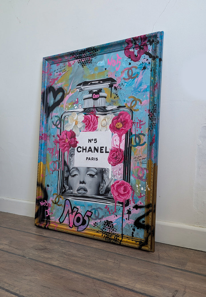 "Marilyn Monroe X Chanel No.5" – a bold embellished art print by Ian Salmon, featuring a black-and-white portrait of Marilyn Monroe embedded in a painted Chanel No.5 bottle, surrounded by vivid pink and red Camellia flowers, graffiti-style branding, and set against a turquoise, pastel blue, and gold accented background with a hand-painted custom frame.