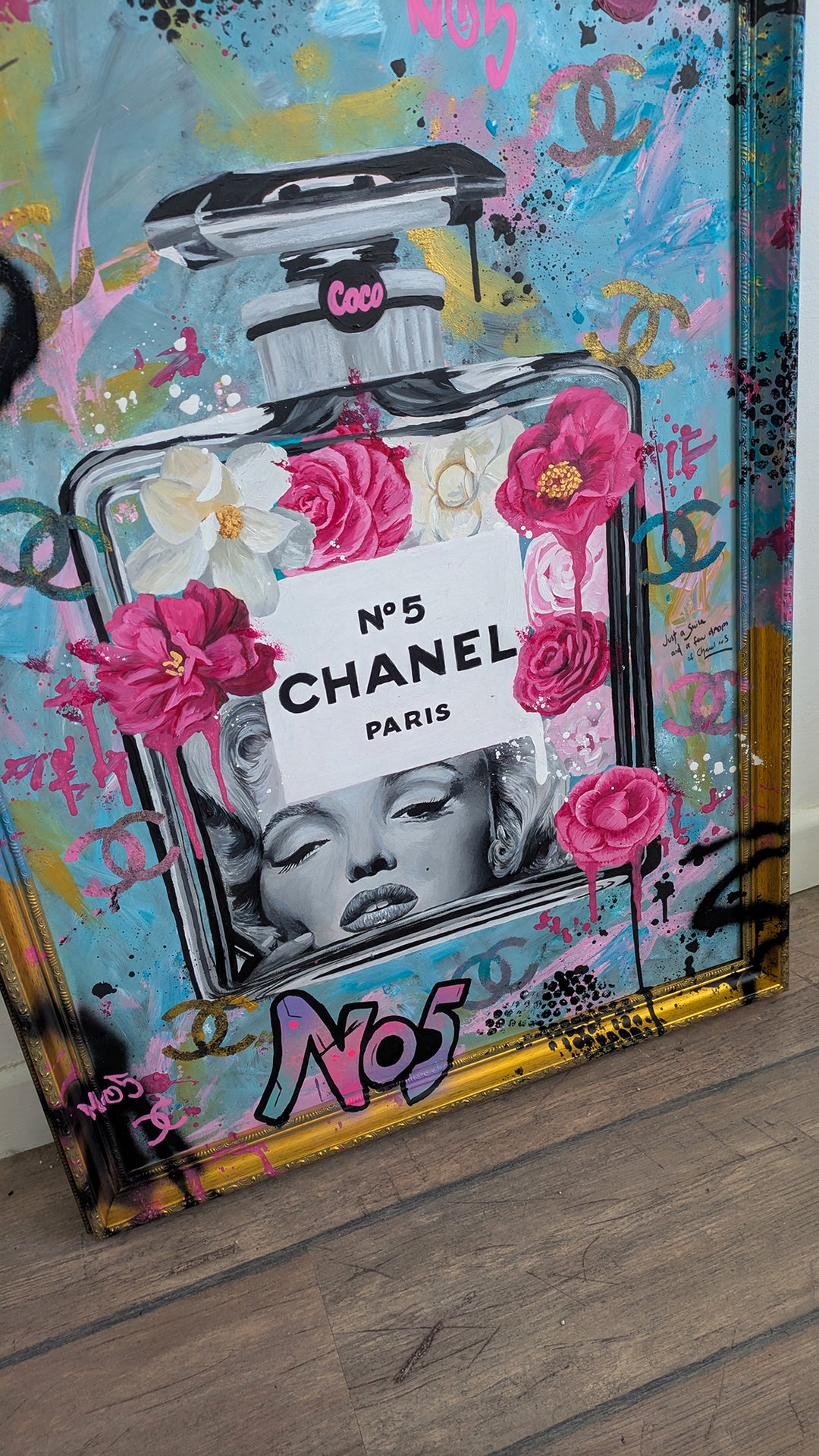 "Marilyn Monroe X Chanel No.5" – a bold embellished art print by Ian Salmon, featuring a black-and-white portrait of Marilyn Monroe embedded in a painted Chanel No.5 bottle, surrounded by vivid pink and red Camellia flowers, graffiti-style branding, and set against a turquoise, pastel blue, and gold accented background with a hand-painted custom frame.