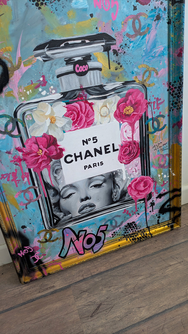 "Marilyn Monroe X Chanel No.5" – a bold embellished art print by Ian Salmon, featuring a black-and-white portrait of Marilyn Monroe embedded in a painted Chanel No.5 bottle, surrounded by vivid pink and red Camellia flowers, graffiti-style branding, and set against a turquoise, pastel blue, and gold accented background with a hand-painted custom frame.