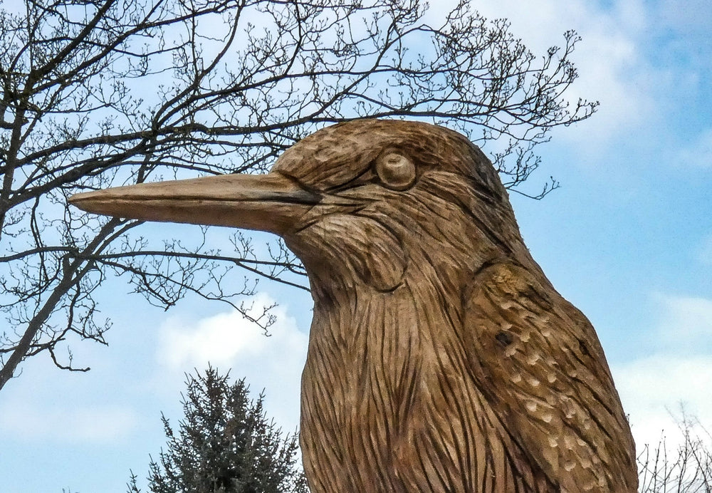 Large hand-carved wooden sculpture of a kingfisher perched on a tree stump, detailed with fine feather textures and set against a garden backdrop.