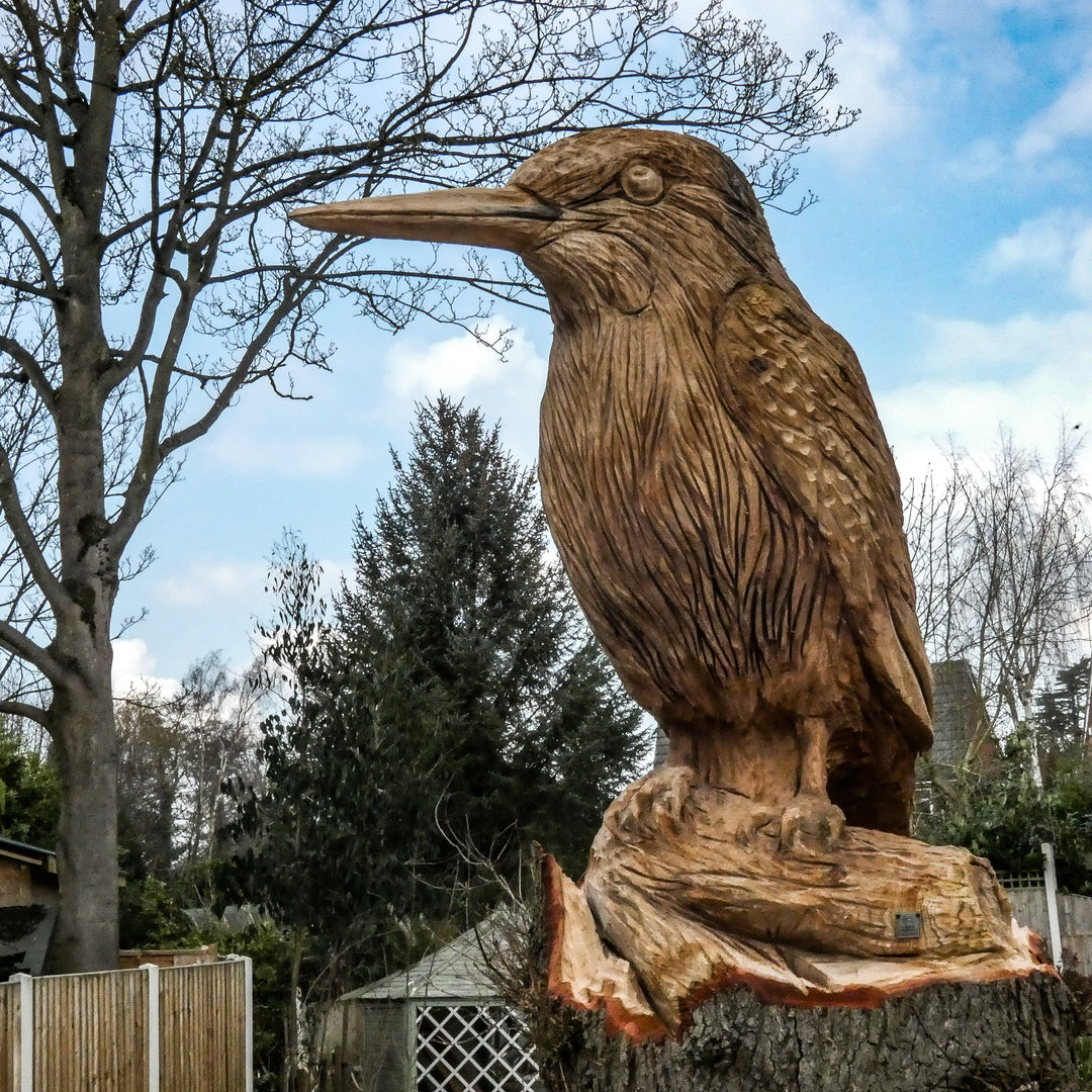 Large hand-carved wooden sculpture of a kingfisher perched on a tree stump, detailed with fine feather textures and set against a garden backdrop.