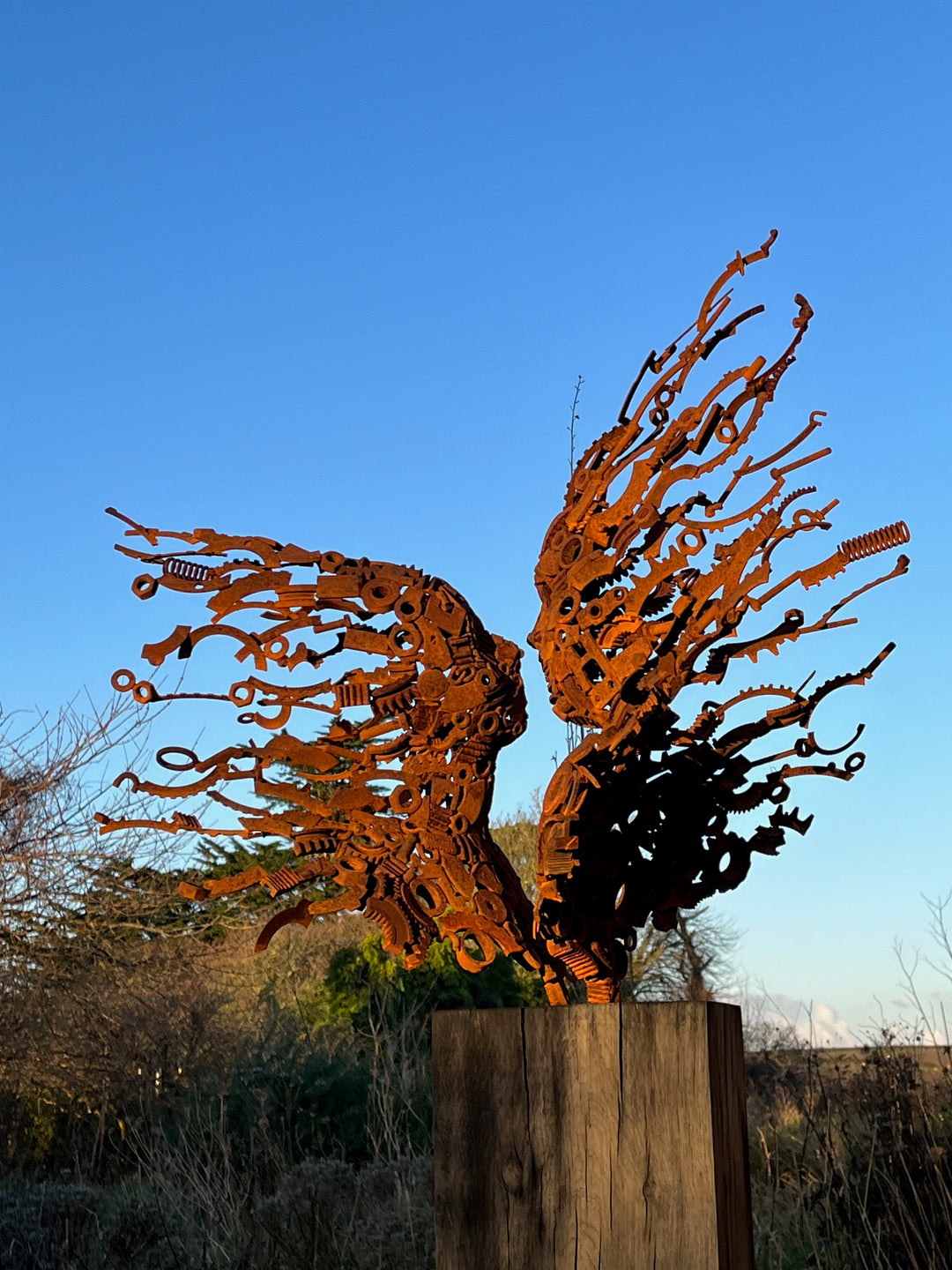 KISMET by Penny Hardy — a limited edition sculpture crafted from recycled metal, depicting two wind-blown figures leaning in to kiss, their bodies and hair formed from intricate mechanical parts and rusted fragments, mounted on a timber plinth in an open landscape.
