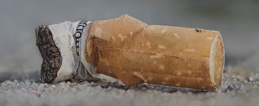 Simon Taylor, Kiss Me Goodbye, oil and acrylic on aluminium, 25 x 60cm – photorealist painting of a discarded cigarette butt lying on gritty ground, with detailed ash, torn paper and shallow depth of field in muted grey and brown tones.